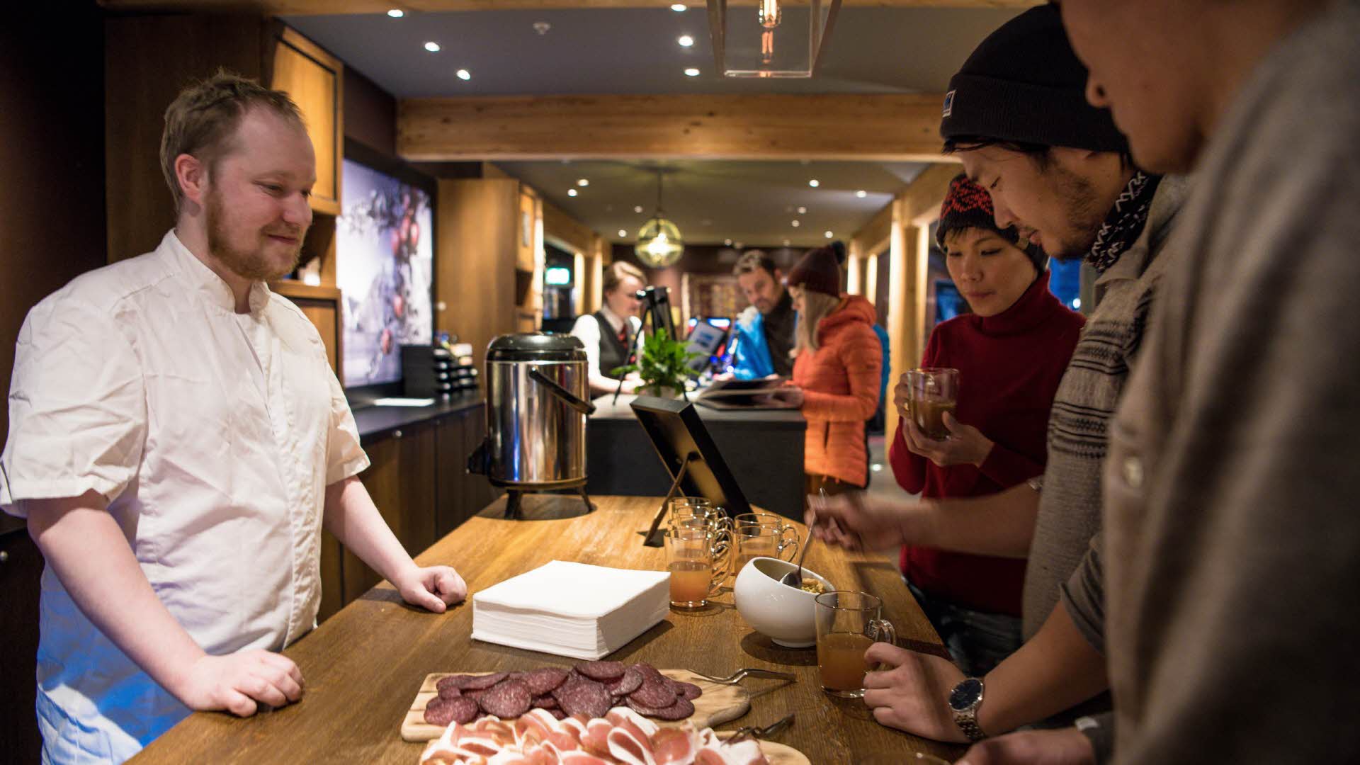 A chef and three hotel guests standing by a bar with mulled wine and cured meats. Two other people checking in at the reception in the background.