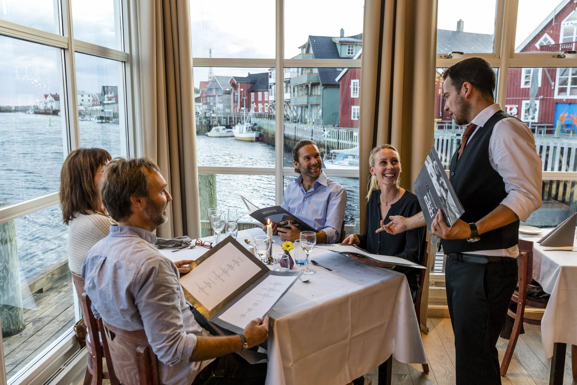 Quatre convives et un serveur à une table du restaurant Blue Fish, à Henningsvær. De grandes fenêtres avec vue sur la jetée.