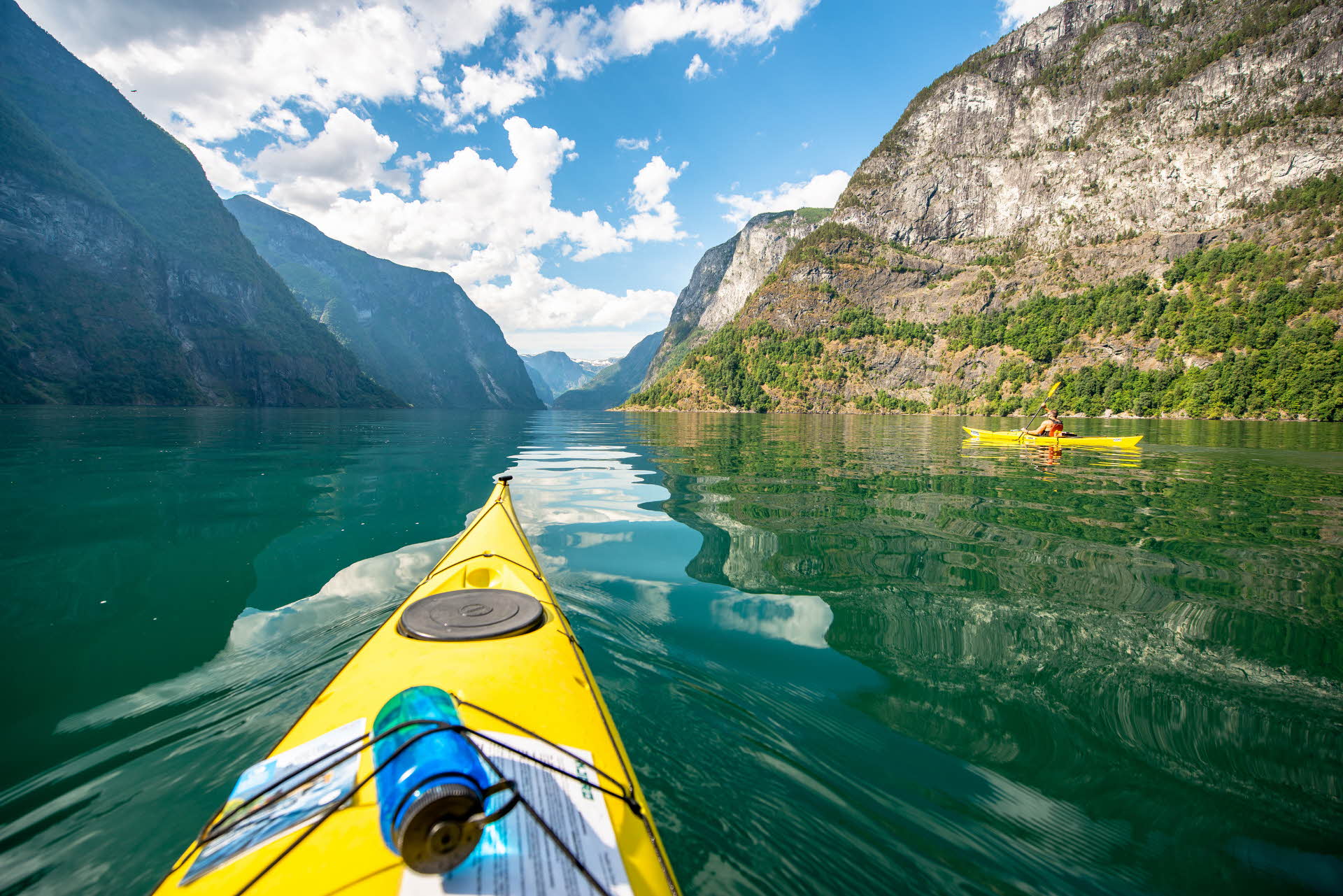 Une moitié d’un kayak jaune au premier plan, une vue d’un autre kayak et des montagnes bordant le fjord.
