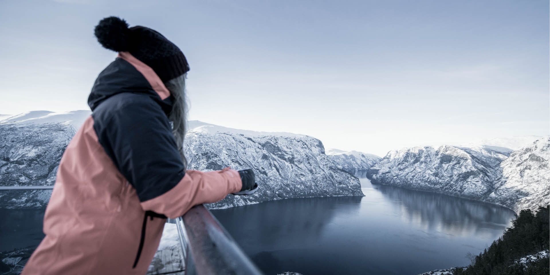 En kvinne står på Stegastein Utsiktpunkt og ser utover Aurlandsfjorden og det snødekte UNESCO verdensarvlandskapet. 