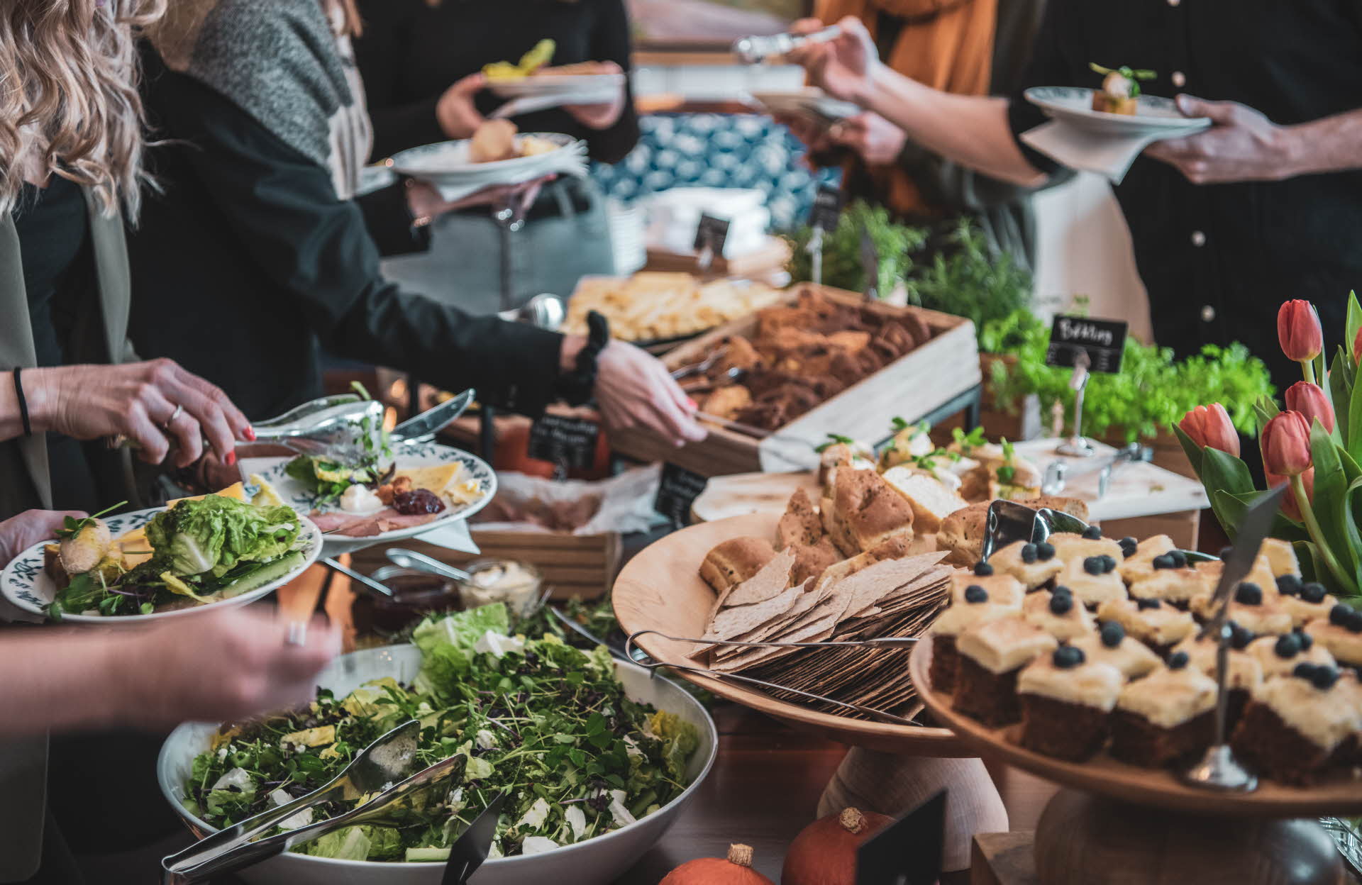 Les mains de plusieurs personnes se servant à un buffet composé de feuilles de salade, de gâteau à la carotte, de pain et de viande séchée, à Flåm