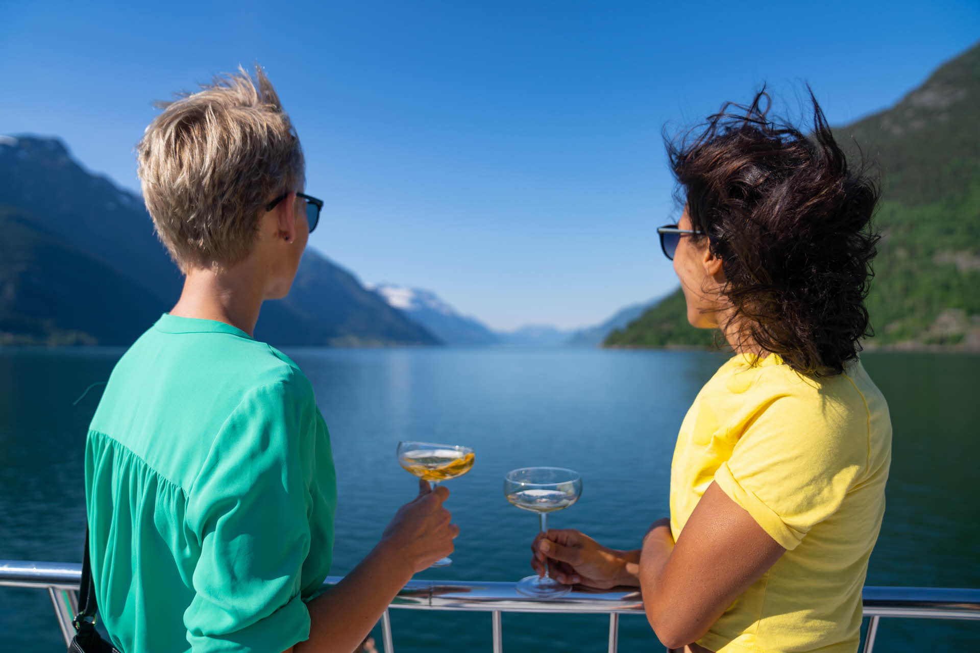 Two women in bright colours with cider glasses in their hands look out on the Hardangerfjord on a fjord cruise