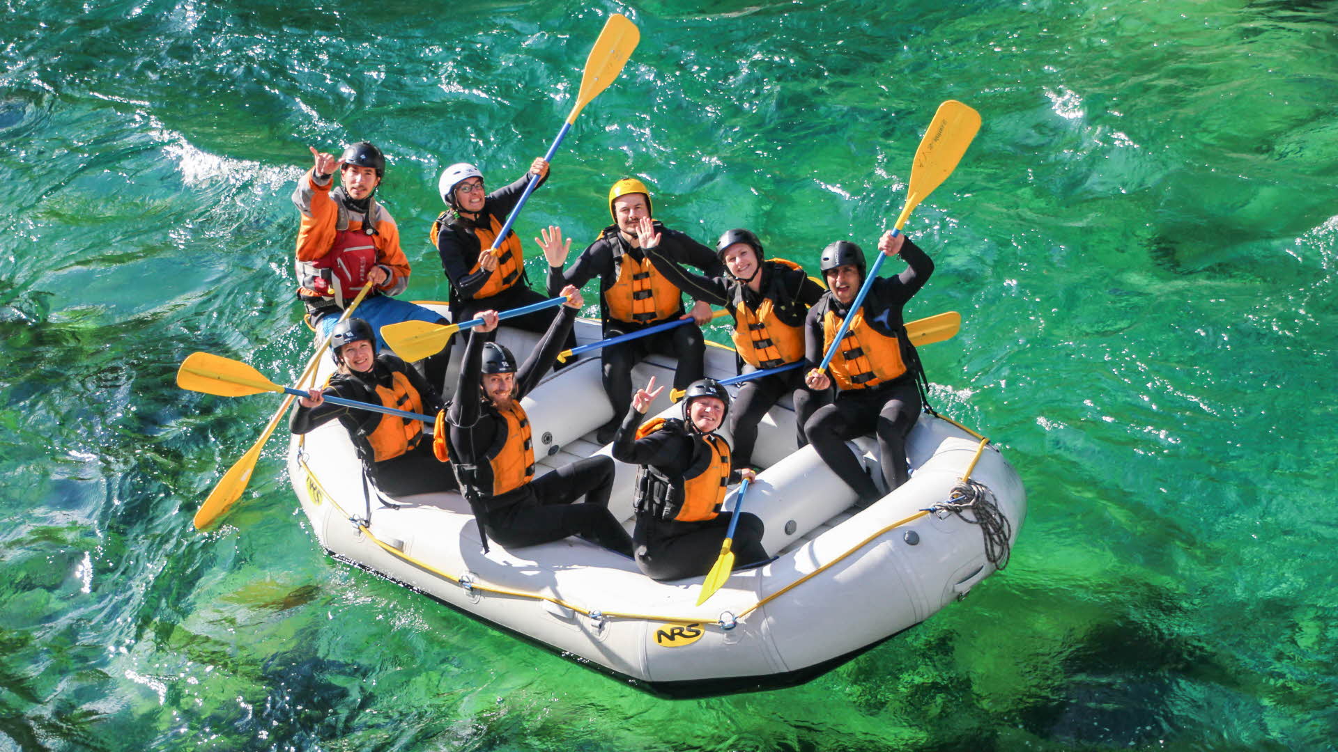 A raft with eight people waving their hands on a clear green river.