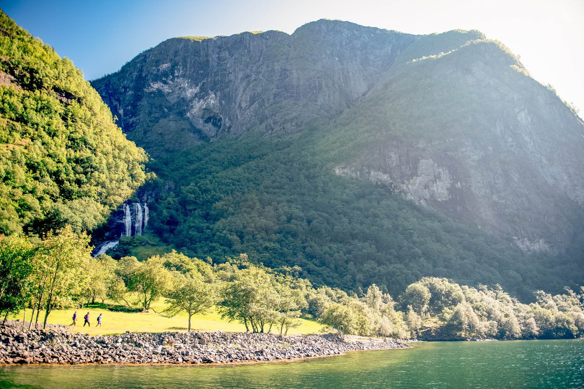Three walkers in a green field under tall mountains and a waterfall. Nærøyfjord in the foreground. 