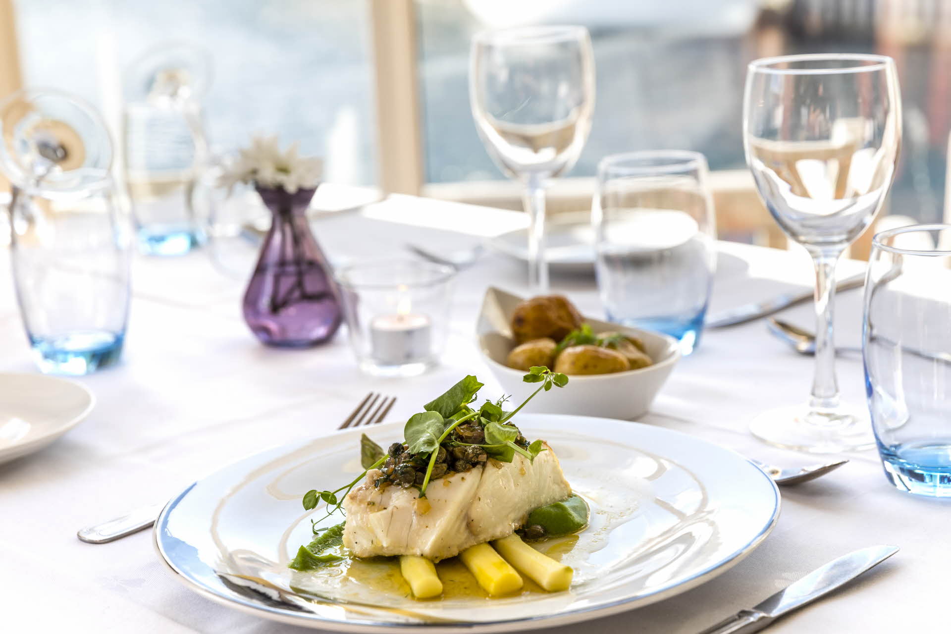 En tallerken med fisk og tilbehør på et dekket bord. Skål med poteter, glass og vindu. 