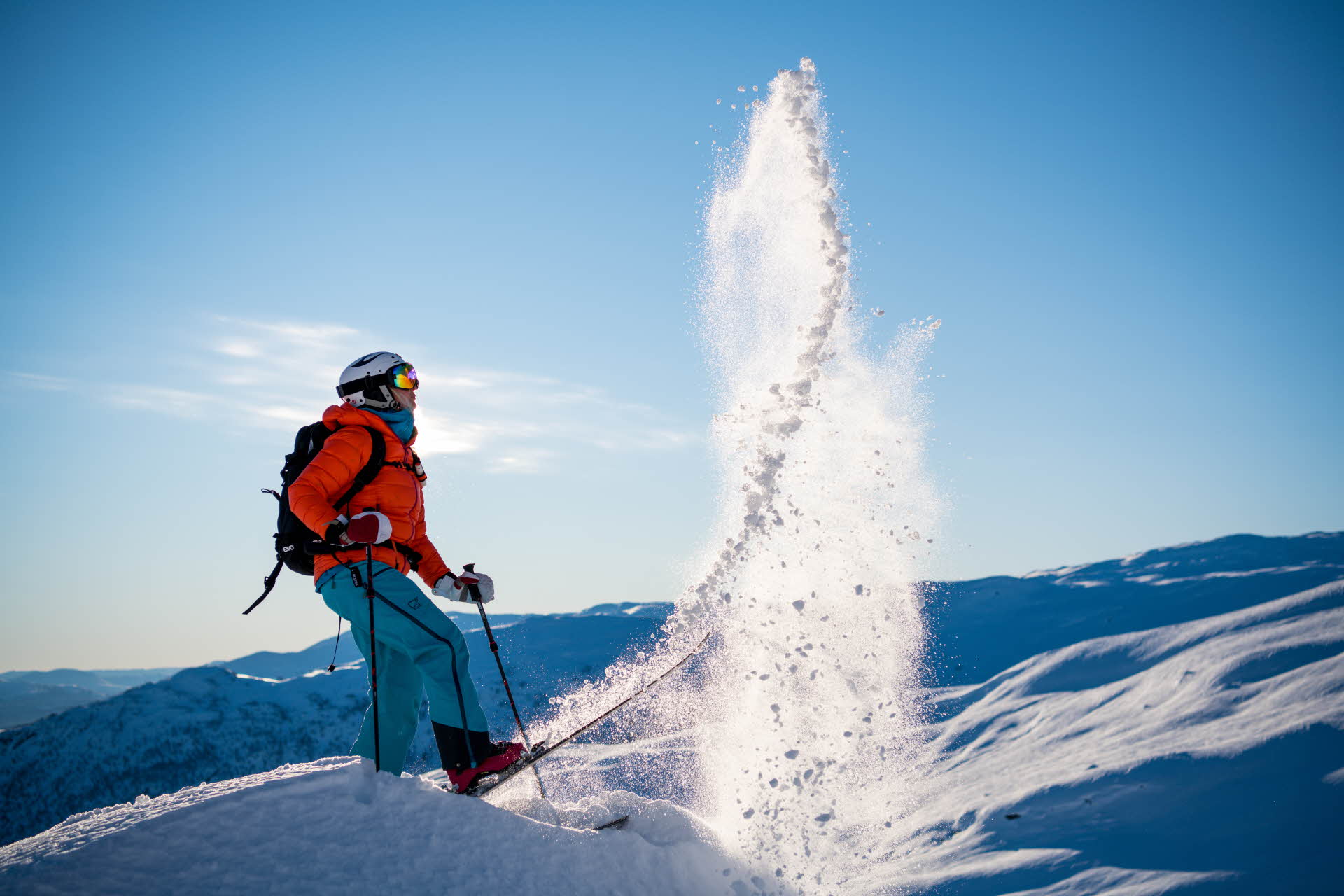 A skier in a red jacket, blue pants and white helmet flips powder snow in the air with a ski on top of a mountain
