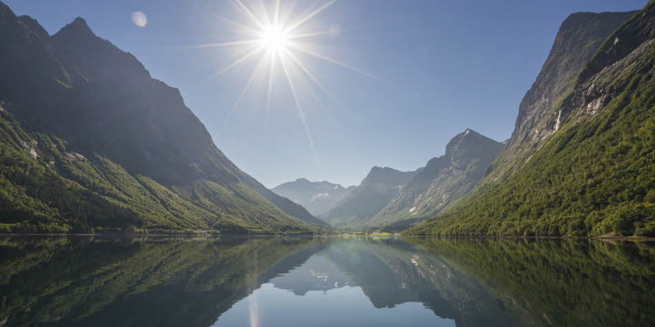 Sun flare as watching the high and dramatic peaks of the Hjorundfjord landscape is mirrored in the calm and crystal-clear sea