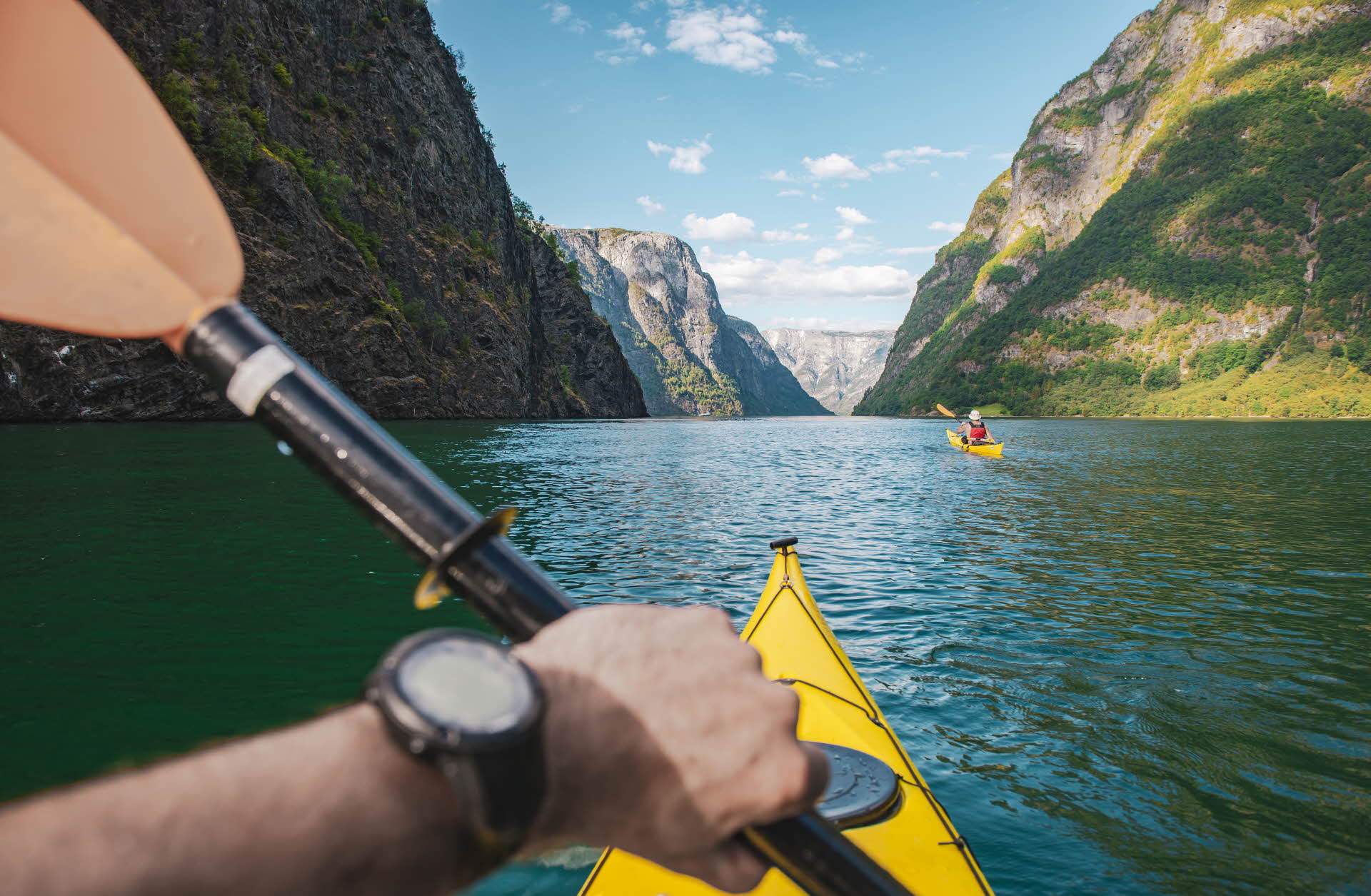 A hand holding a paddle over a yellow kayak. Another kayaker, the Nærøøyfjord and mountains in the foreground.