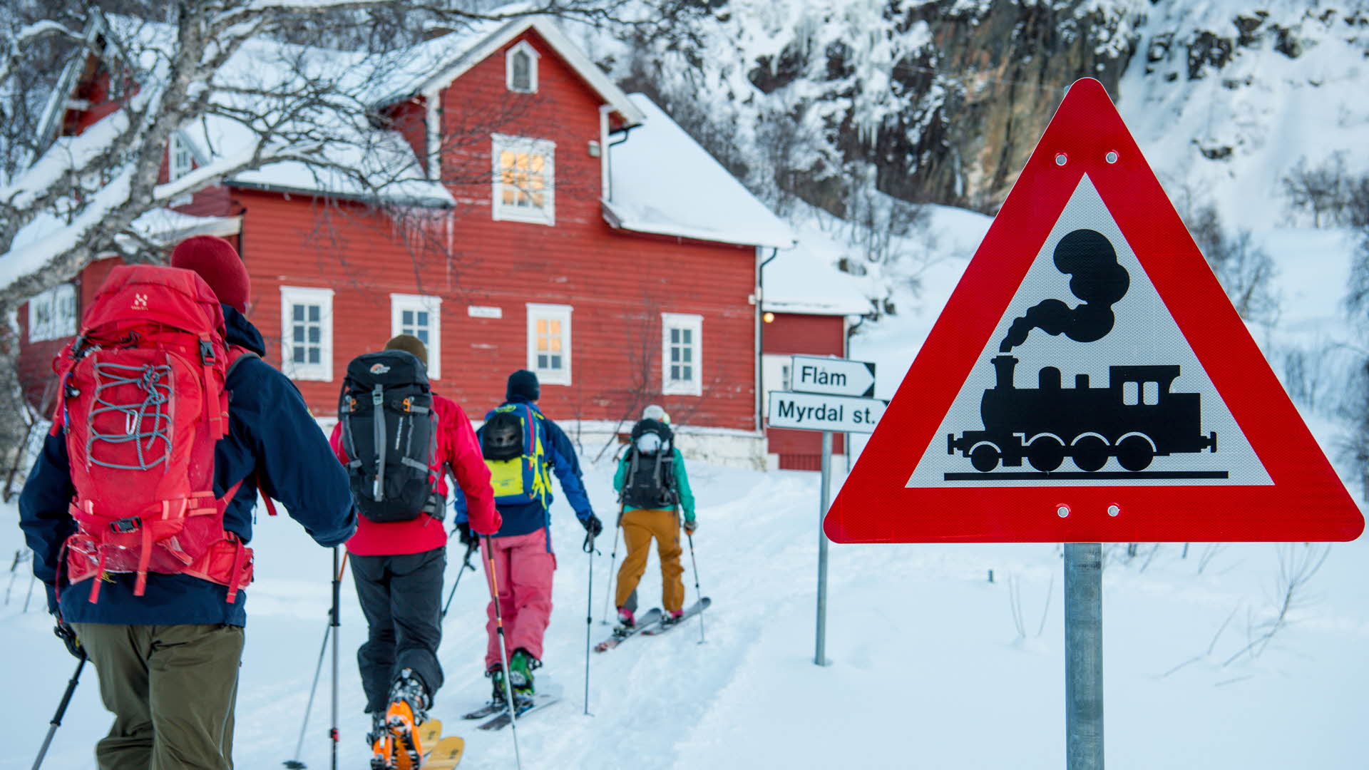 Quatre skieurs de randonnée en route vers la maison d’hôtes rouge de Vatnahalsen passant devant le panneau signalant le chemin de fer de Flam en hiver