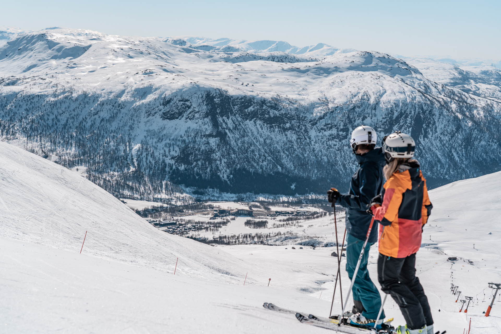 Dos esquiadores en la parte alta de una pista mirando hacia Myrkdalen Mountain Resort y las montañas alrededor.