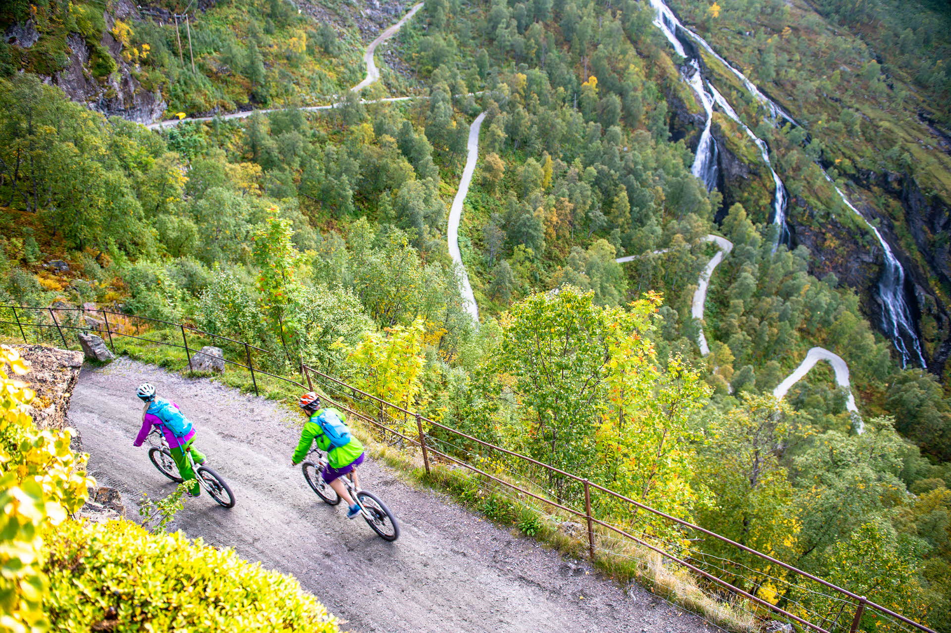 Two people on bikes cycling along a gravel road. Hairpin bends and waterfall below. 