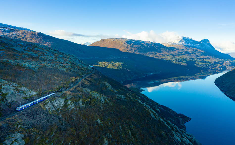 Le train de l’Arctique passant devant une petite cabane rouge sur la ligne d’Ofoten, en direction de Narvik, avec le Rombaksfjord et les montagnes en arrière-plan