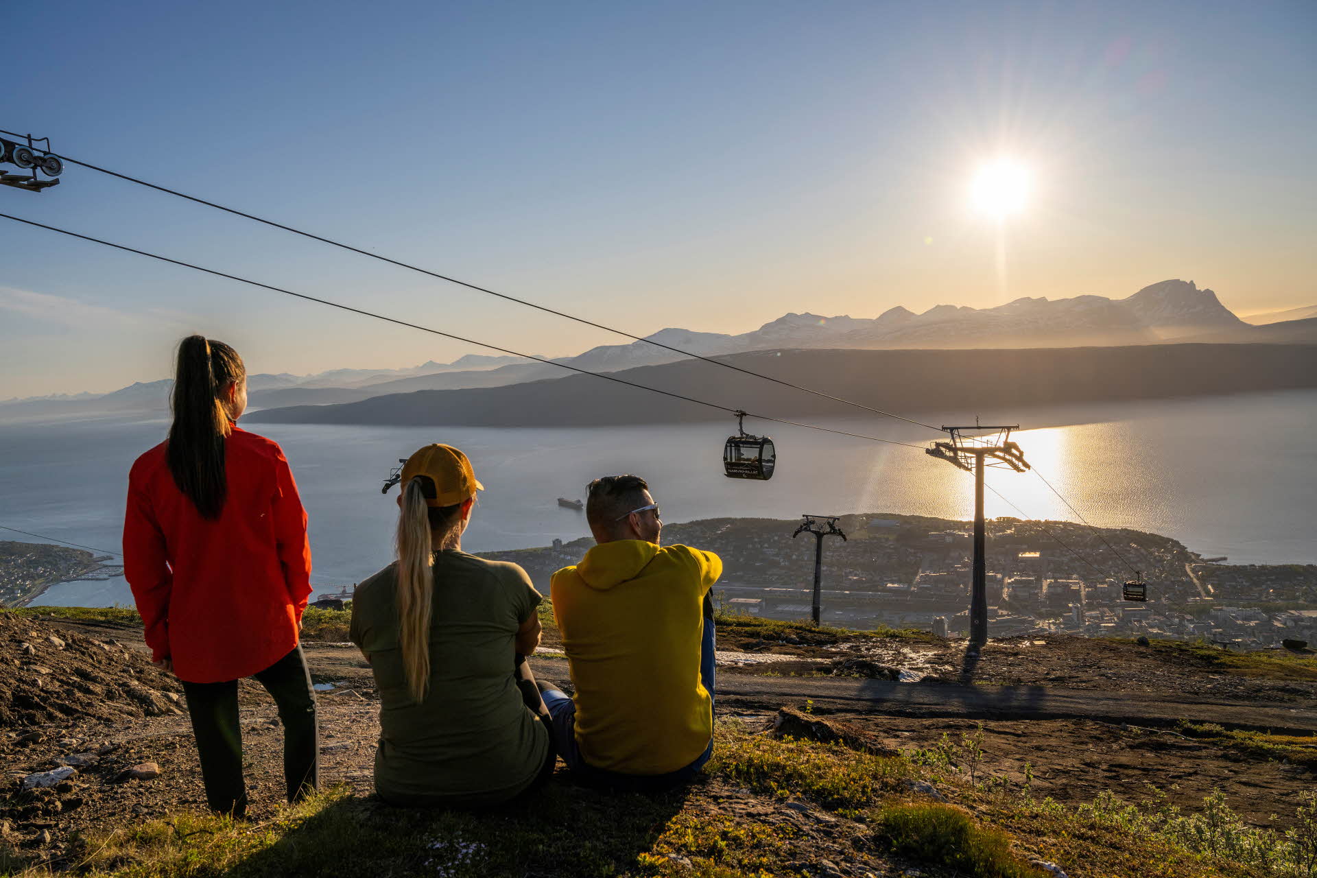 Tres personas mirando hacia Narvik, el teleférico y el fiordo con islas montañosas en el horizonte.