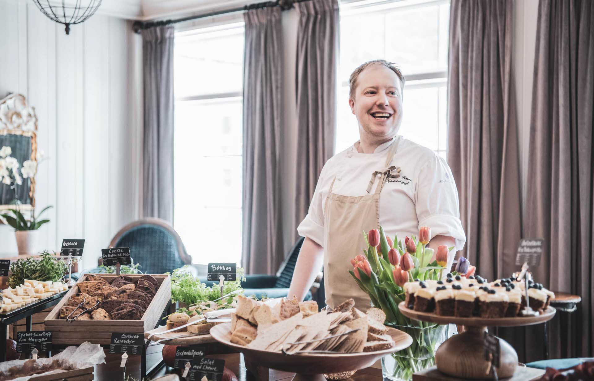 A chef behind a buffet of small dishes incl. cake, flatbread and cheese.