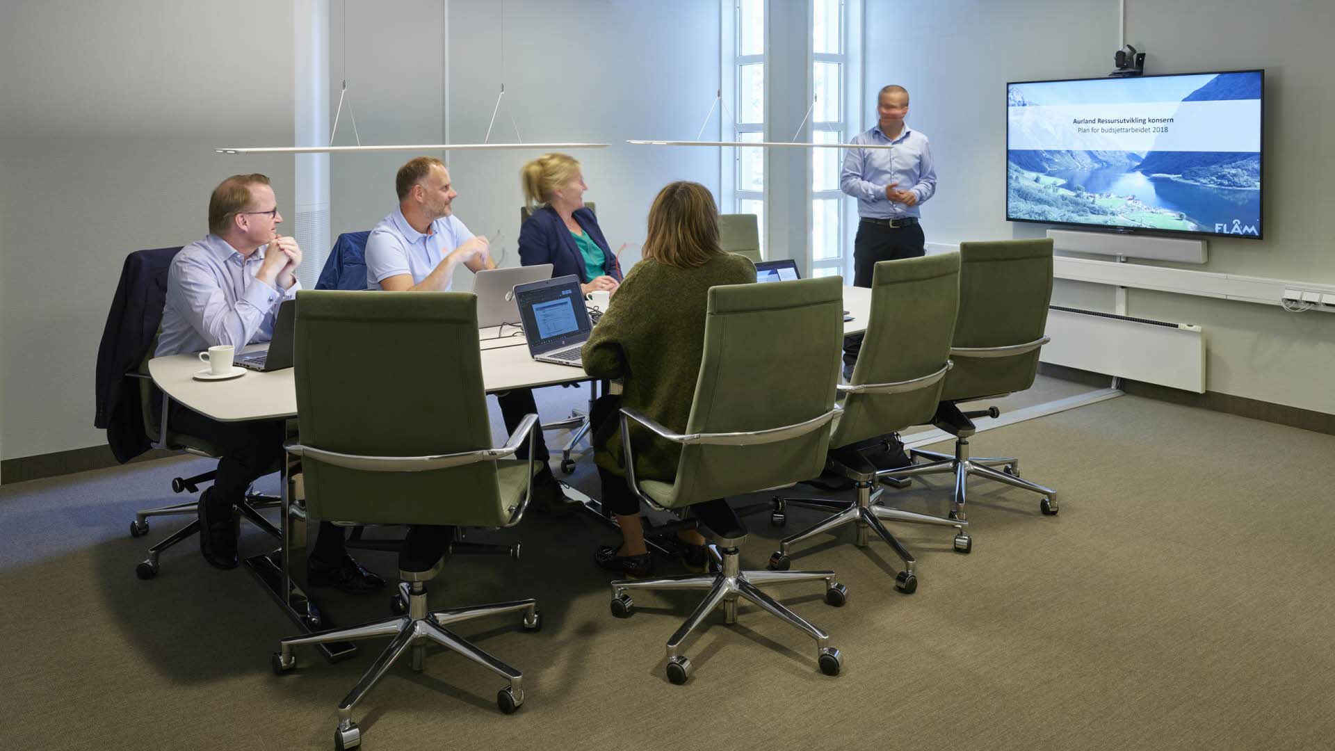 Four people around a conference table watching a man making a presentation on a TV screen.
